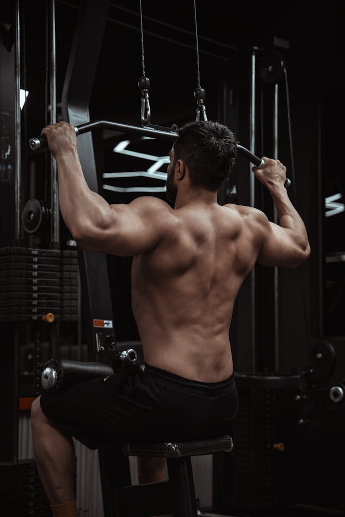 who-we-are Muscular man performing a lat pulldown exercise in a dimly lit gym for strength training.