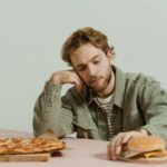 A man sits thoughtfully at a table with a pizza and burger in a minimalist indoor setting.